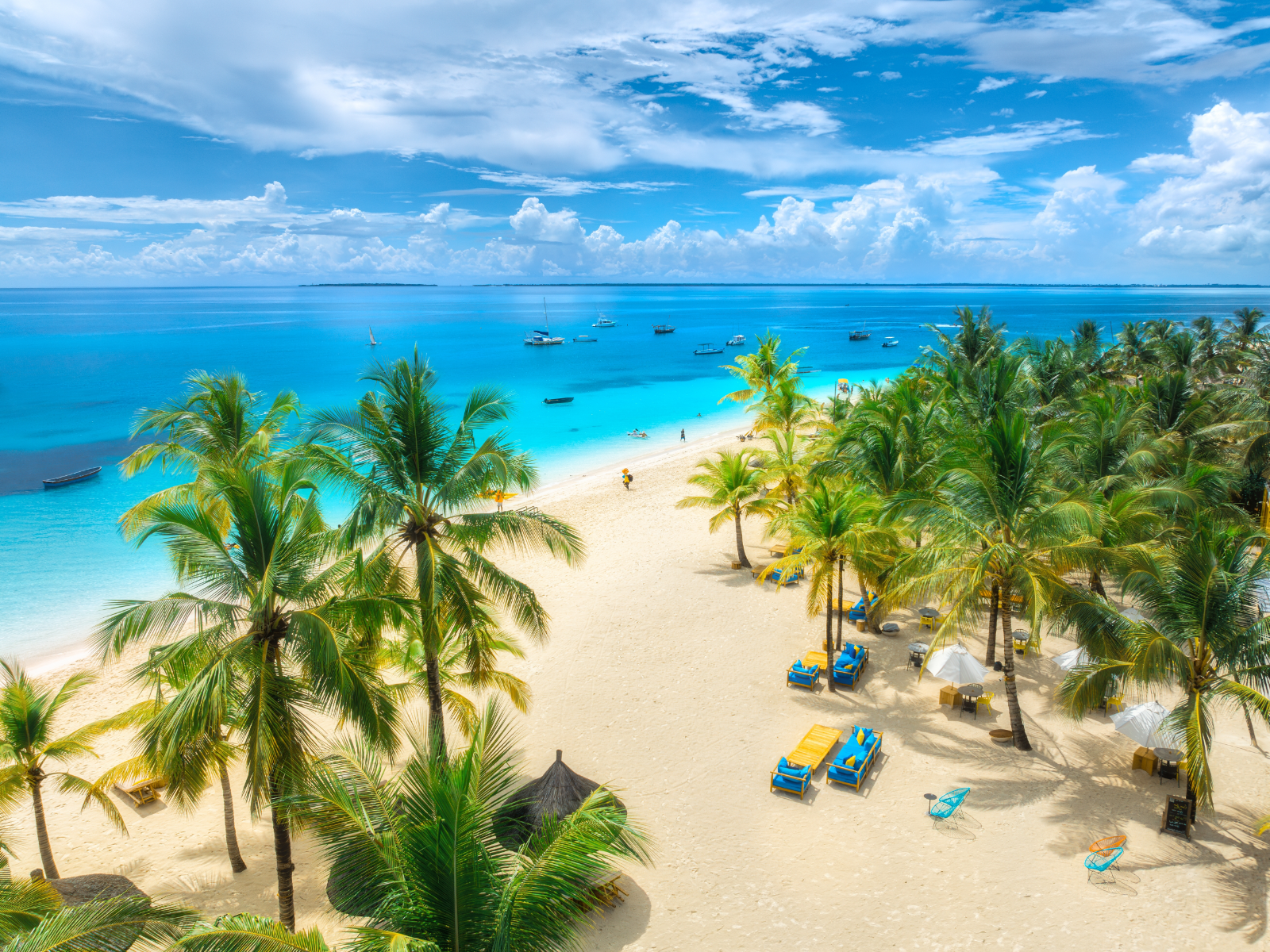 Aerial view of a white sand beach with palm trees, sunbeds, umbrellas, and yachts along a turquoise ocean at sunset